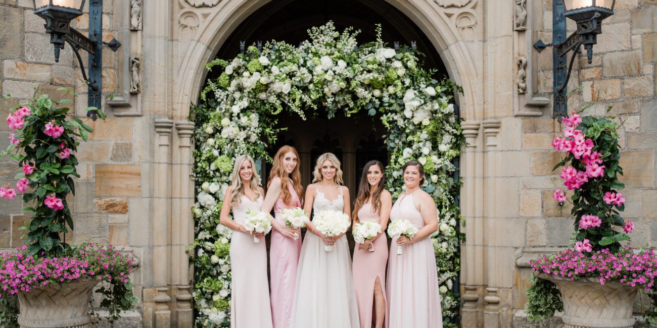 Bride and Bridesmaids pose outside Meadow Brook Hall's front gates, decorated with a flower arch.
