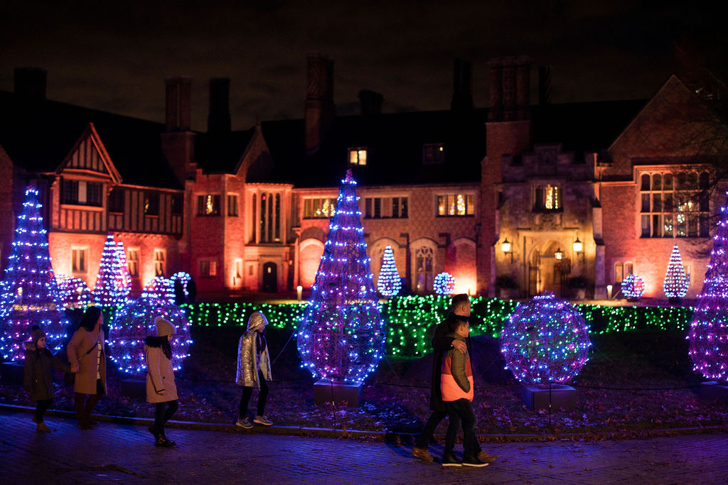 A group of adults and children walk past brilliantly lighted decorations with Meadow Brook Hall in the background